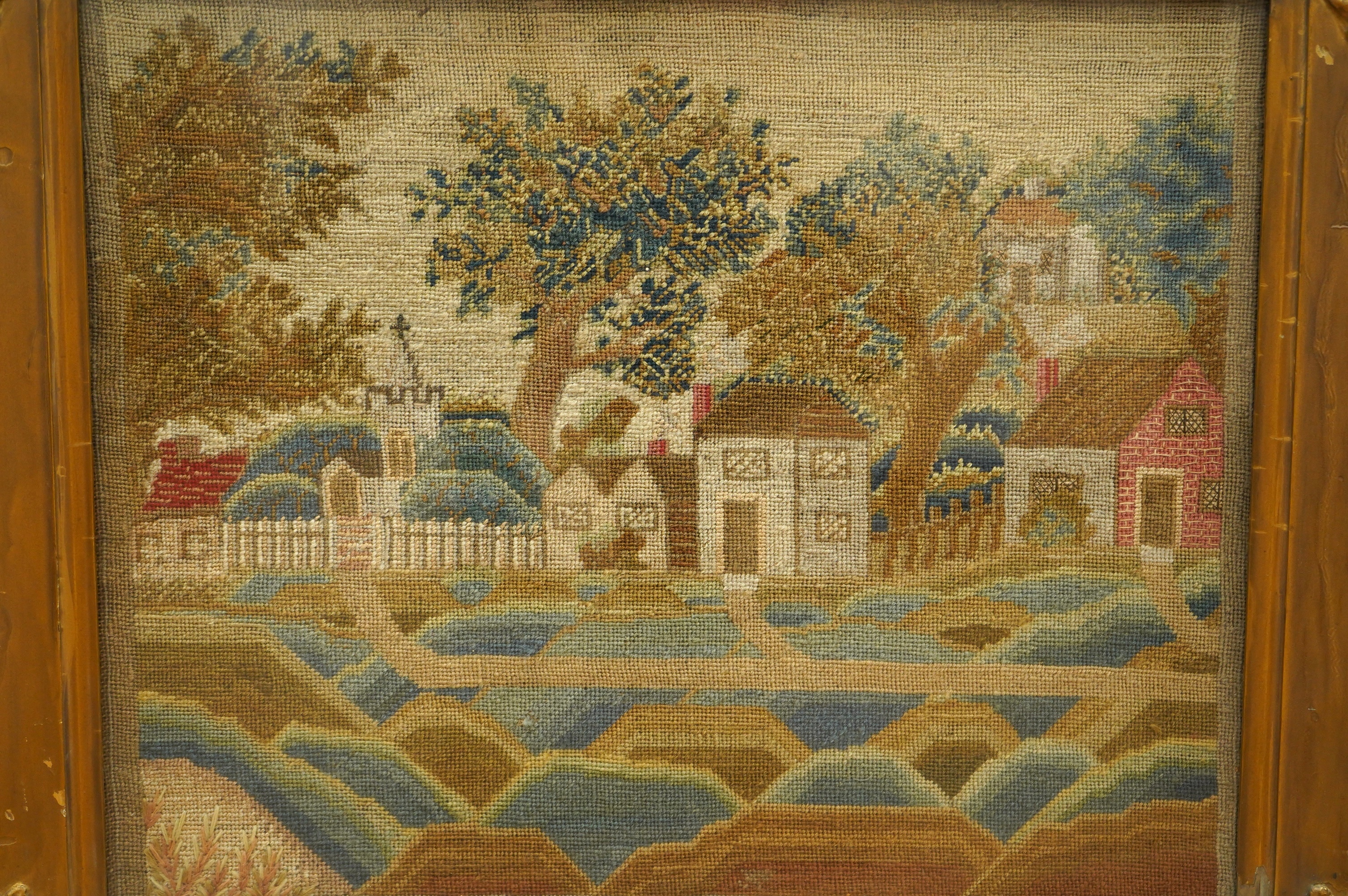 A framed 19th century woolwork of a village scene of cottages, houses, a church amongst trees and a picket fence with paths leading to another, within an undulating foreground, the leaves of the trees worked in various s